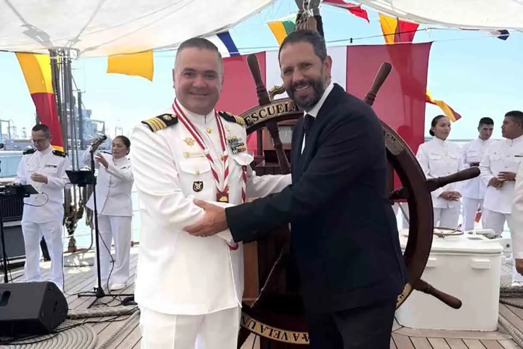 Dos hombres en un evento formal en un barco, uno con uniforme naval y conmedallas, saludando a otro en traje oscuro. Al fondo, otros miembros de la tripulación y decoración náutica.