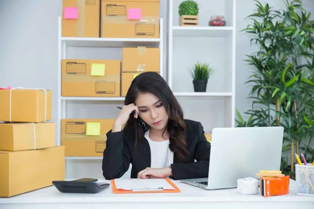 Mujer de negocios pensativa sentada frente a una computadora portátil en una oficina con cajas de envío y notas adhesivas de colores.