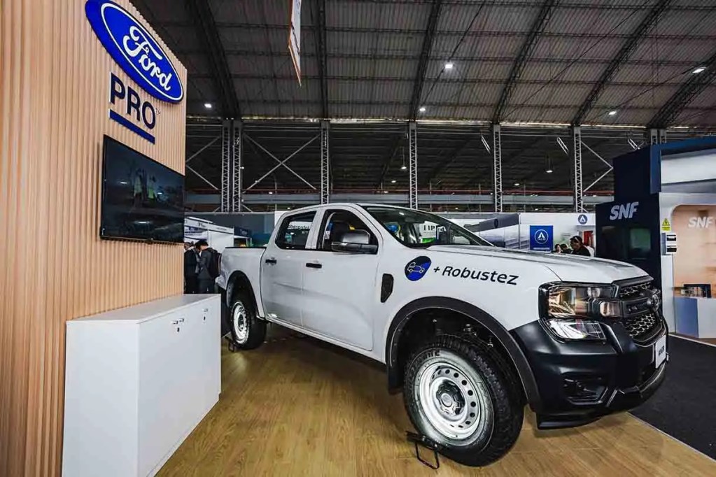 Camioneta Ford Pro blanca exhibida en un evento, con un fondo de un stand de presentación y una pantalla de televisión.