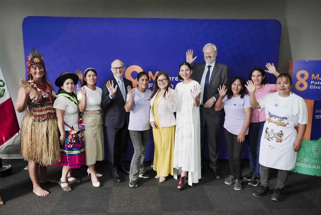 Grupo de mujeres de diversas culturas y hombres posando juntos, saludando, con bandera de México al fondo y fondo publicitario en el evento 'Futuro Circular'.