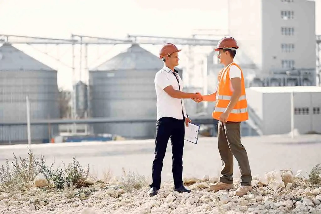 Dos hombres se dan la mano en un sitio de construcción, uno lleva casco y chaleco reflectante.