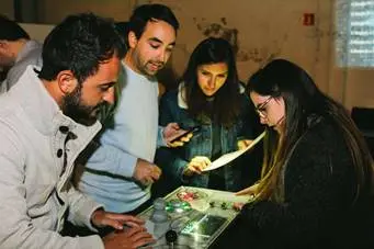 Cuatro personas interactuando con una mesa de trabajo, revisando documentos y dispositivos electrónicos en un entorno colaborativo.