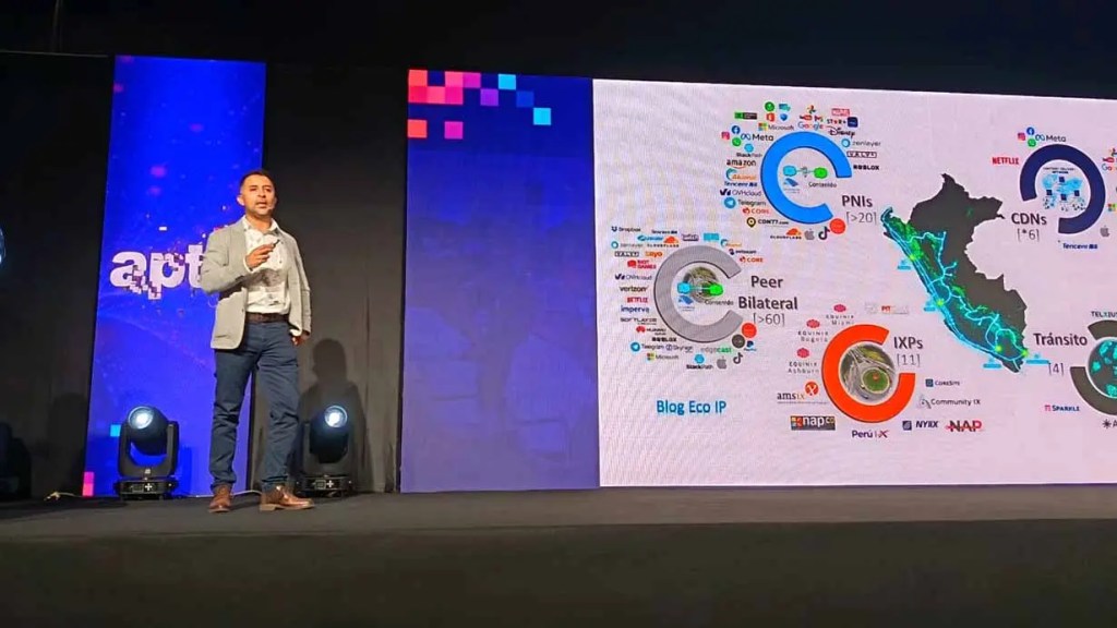Presentador en un escenario durante una conferencia, con una diapositiva que muestra un mapa de interconexiones de red en América del Sur.