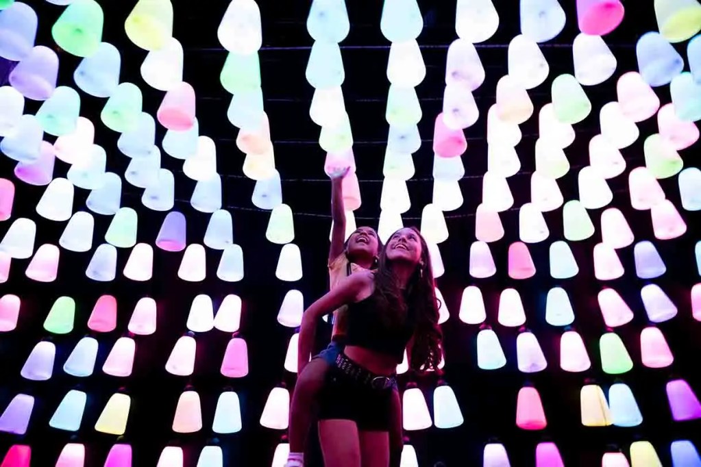 Dos mujeres posando sonrientes bajo una instalación luminosa con luces de colores vibrantes en un fondo oscuro.