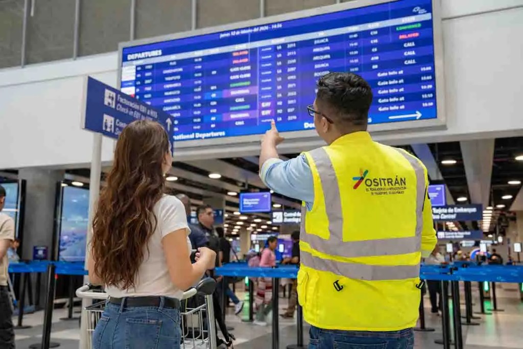 Un hombre en un chaleco amarillo señala una pantalla de salidas en un aeropuerto mientras una mujer lo observa con una maleta.