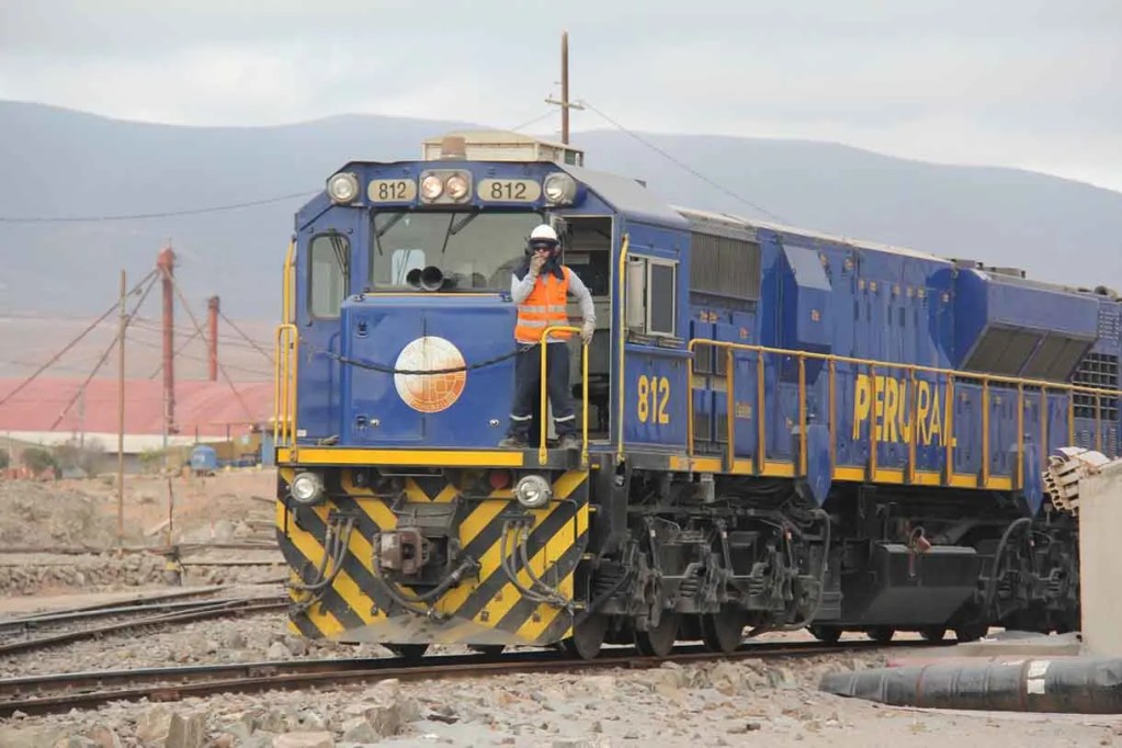 Locomotora de tren 812 de color azul y amarillo, con un conductor a bordo, en un entorno industrial.