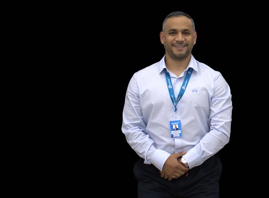 Hombre de pie con camisa de manga larga azul, sonriendo y con una identificación colgante alrededor del cuello, frente a un fondo negro.