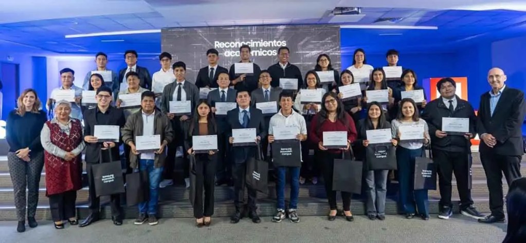 Grupo de personas posando para una foto en un evento de reconocimientos académicos, sosteniendo certificados y bolsas de regalo. Fondo con pantallas que dicen 'Reconocimientos académicos'.