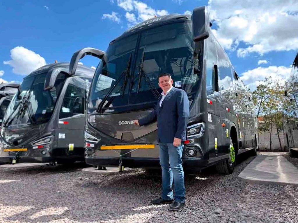 Un hombre de pie junto a un autobús Scania en un aparcamiento al aire libre, con cielo azul y nubes de fondo.