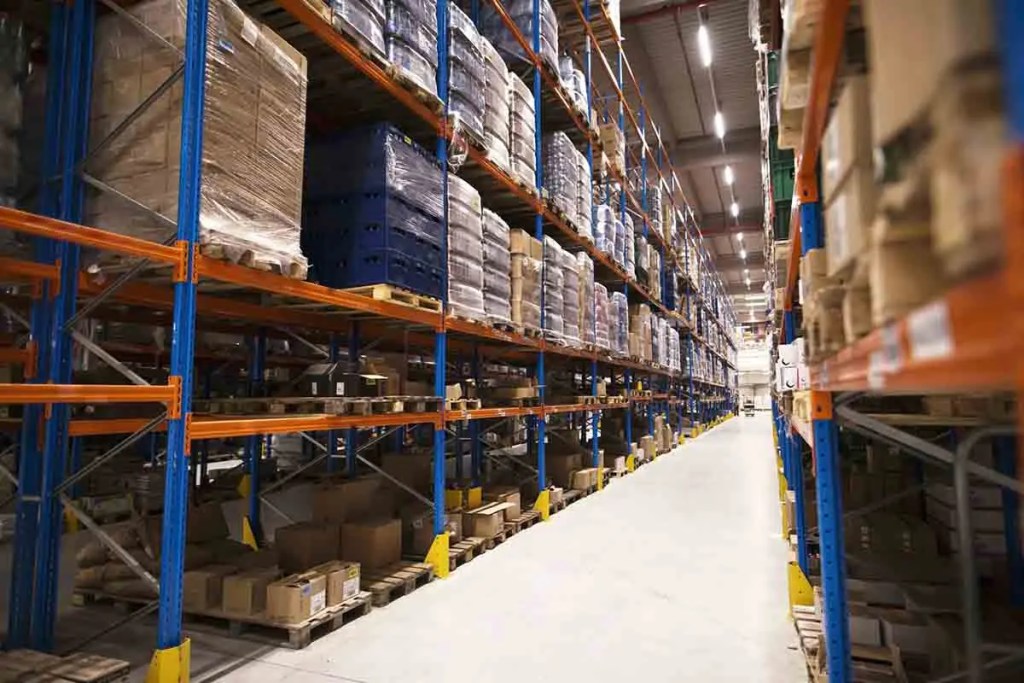 Vista de un almacén con estanterías llenas de cajas y pallets, mostrando un pasillo amplio entre las filas de estanterías