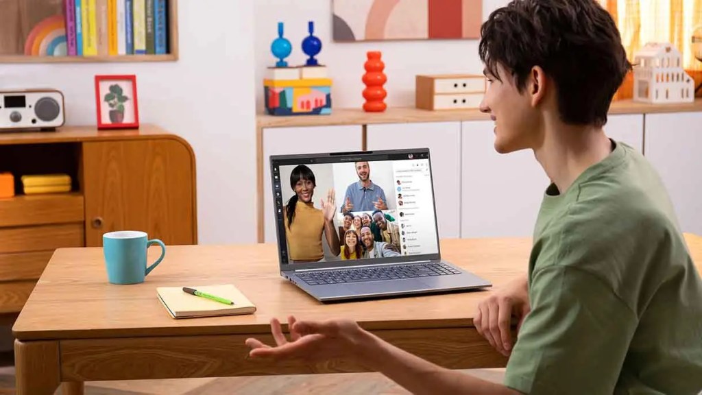 Hombre sentado frente a un laptop con una videollamada abierta, sonríe mientras habla con varias personas en la pantalla. Sobre la mesa hay una taza azul y una libreta con un bolígrafo.