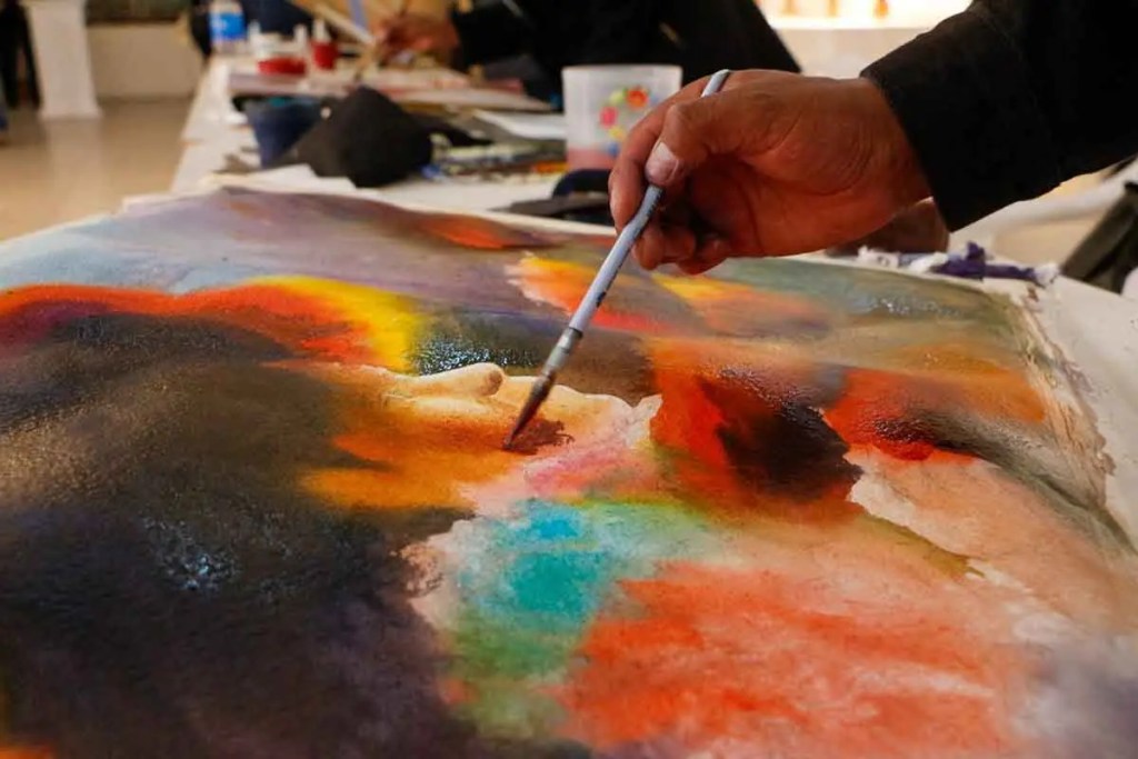 Una mano sostiene un pincel mientras pinta con acuarelas en un papel con colores vibrantes y texturas variadas.