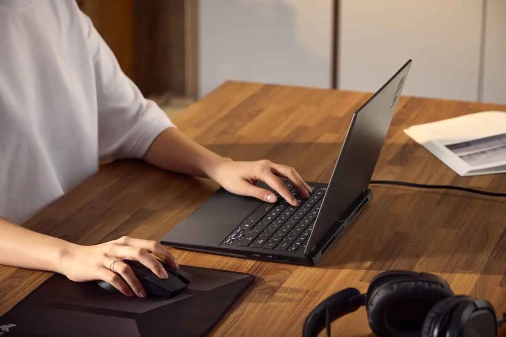 Una persona está usando un portátil en una mesa de madera, sujetando un mouse y con auriculares cerca.