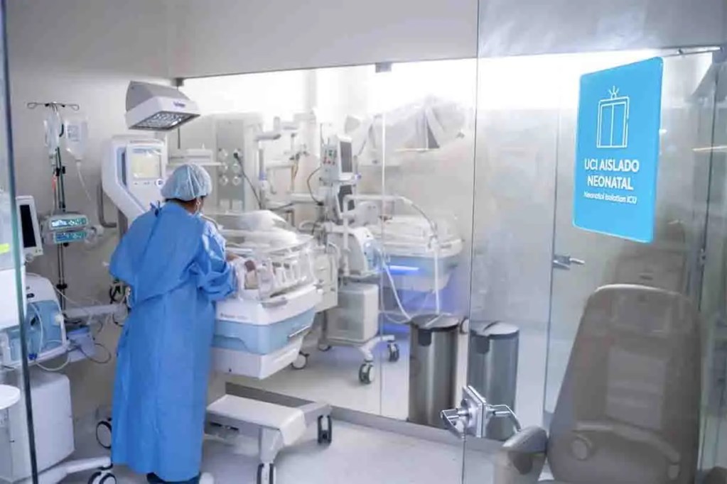 Imagen de una unidad de cuidados intensivos neonatales con un profesional de salud atendiendo un incubadora. Se observa un entorno médico limpio y equipado con tecnología avanzada.