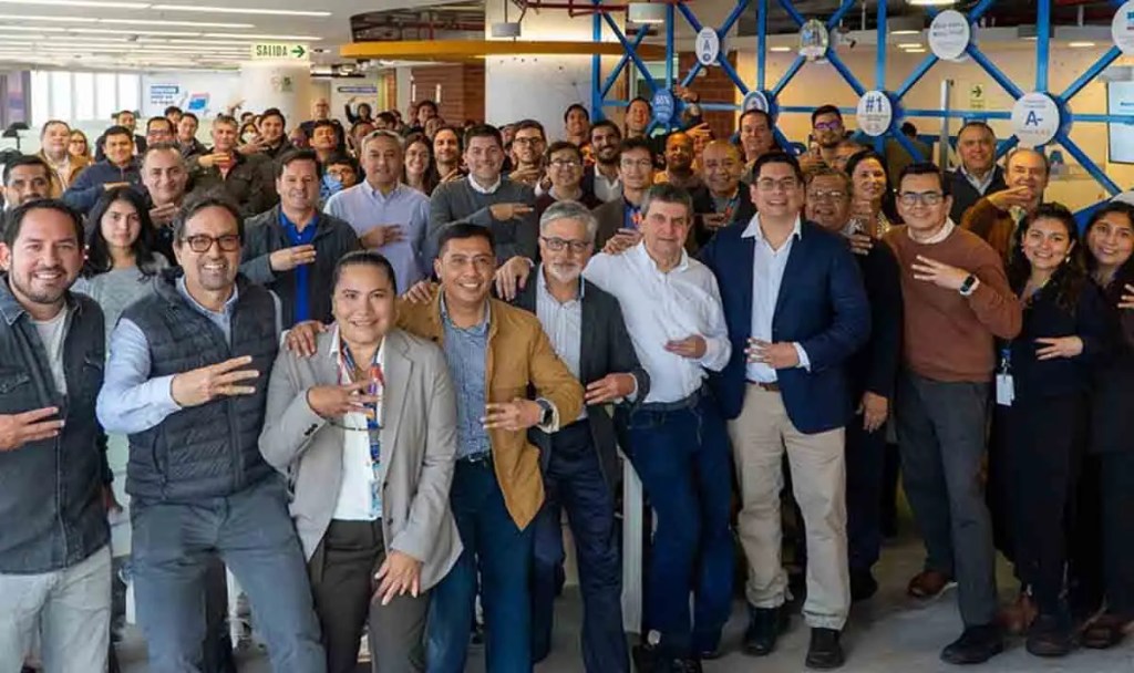 Grupo de personas sonriendo y posando en una oficina, algunos levantando tres dedos en señal de celebración.