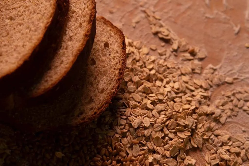 Rebanadas de pan integral apiladas sobre una superficie de madera, con granos de avena esparcidos alrededor.
