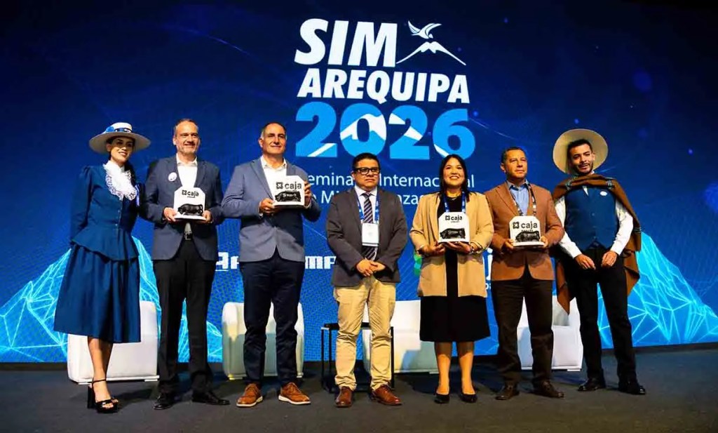 Grupo de personas sonriendo en un escenario durante el evento SIM Arequipa 2026, sosteniendo trofeos y con un fondo azul con motivos tecnológicos.