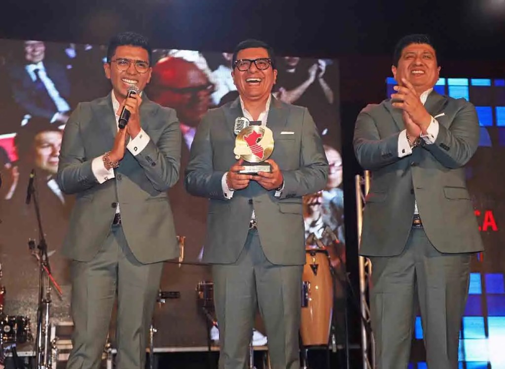 Tres hombres vestidos con trajes grises sonríen y celebran en un escenario, sosteniendo un trofeo. Al fondo se observan multitudes y decoraciones festivas.