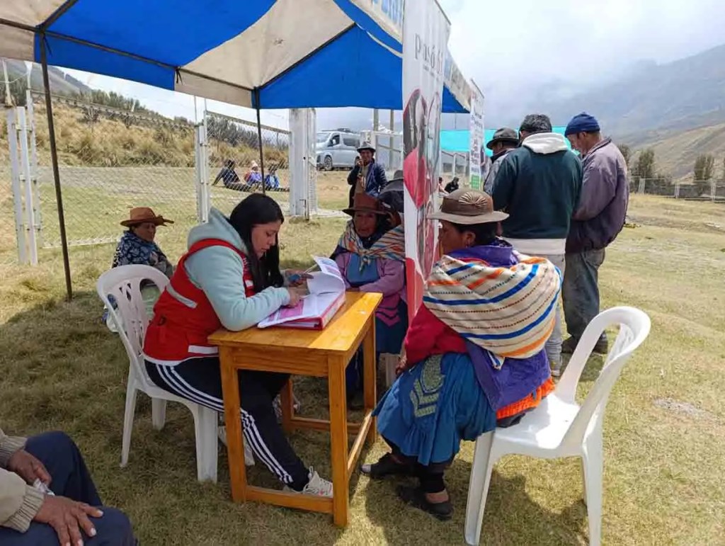 Una mujer con chaleco rojo está escribiendo en una mesa mientras otros se reúnen bajo una carpa en un entorno rural.