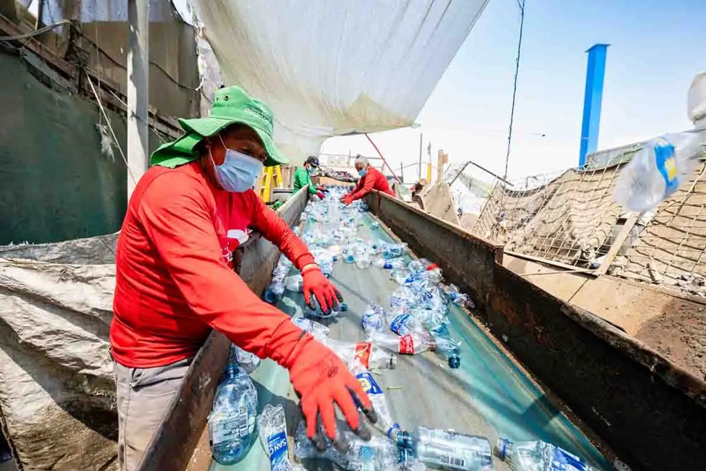 Trabajadores en una planta de reciclaje clasificando botellas de plástico en una cinta transportadora bajo un toldo.