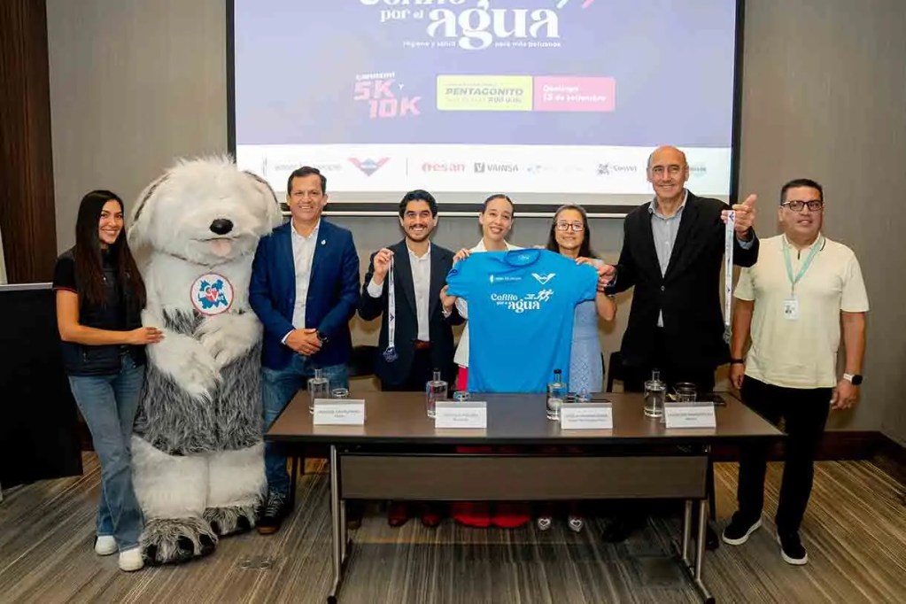 Evento de lanzamiento de la carrera 'Caminando por Agua' con participantes y un personaje de mascota, sosteniendo una camiseta azul y una medalla.