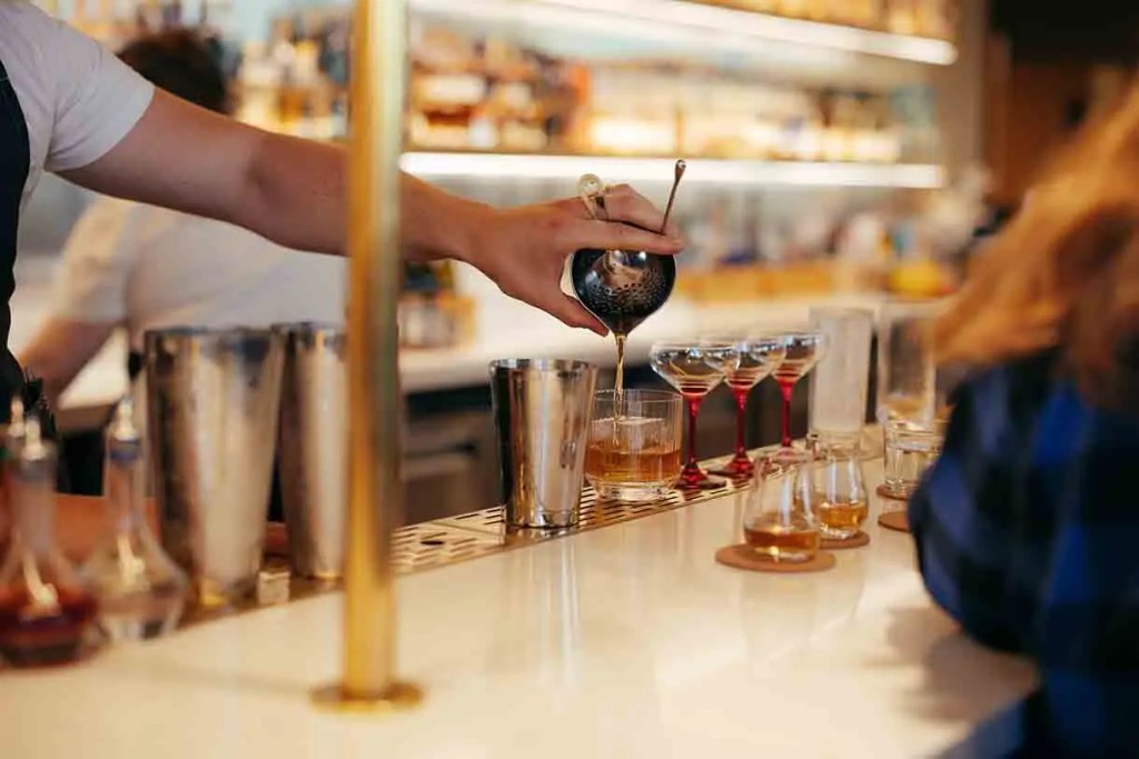 Un bartender sirviendo un cóctel en un bar, con diversas copas y botellas de licor en el fondo.