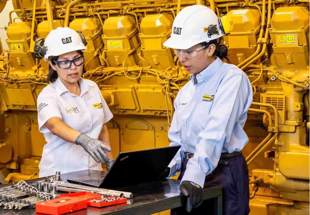Dos mujeres ingenieras en un taller, mirando una computadora portátil con un motor amarillo de fondo. Ambas usan cascos y gafas de seguridad.