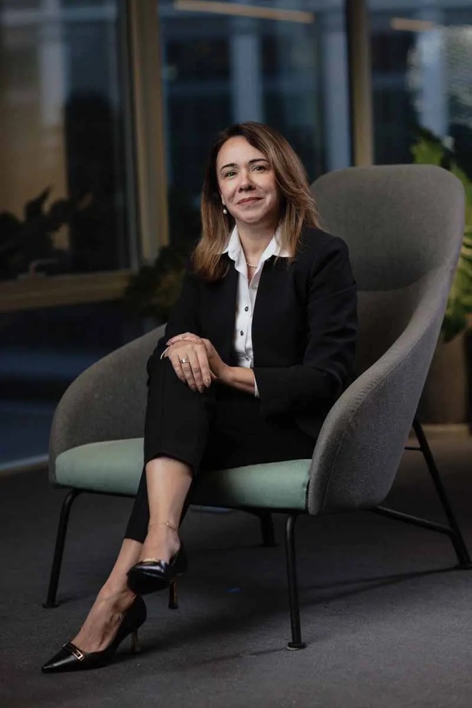 Mujer sentada en un sillón, vestida con traje negro y blusa blanca, sonriendo y con las manos apoyadas en la rodilla.