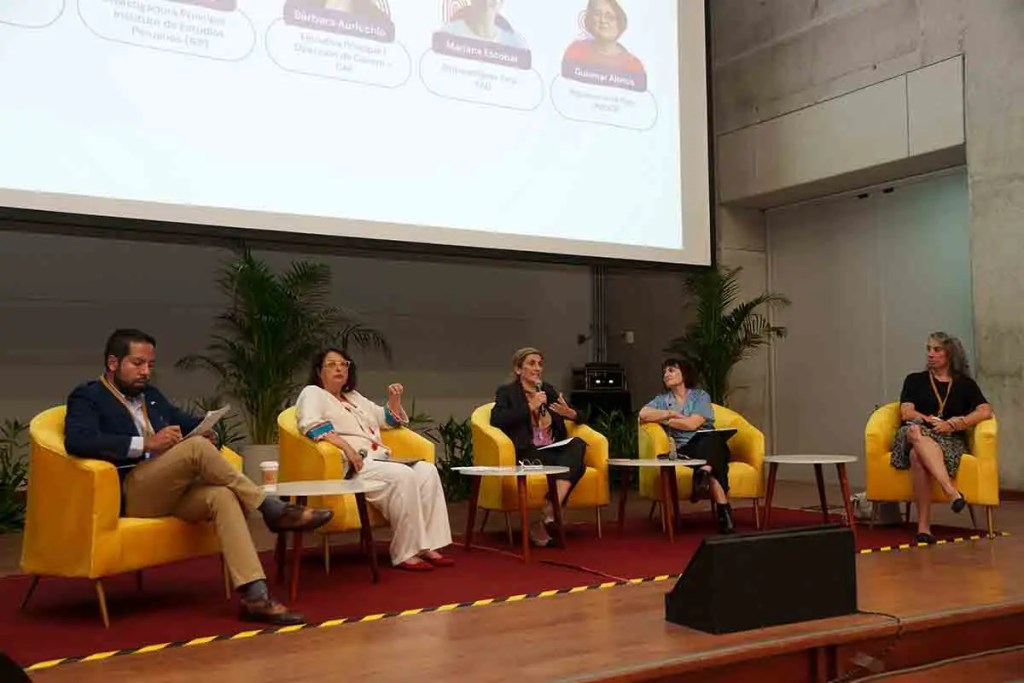 Panel de discusión con cinco participantes sentados en sillas amarillas, en un escenario. Hay plantas alrededor y una pantalla proyectando información detrás de ellos.