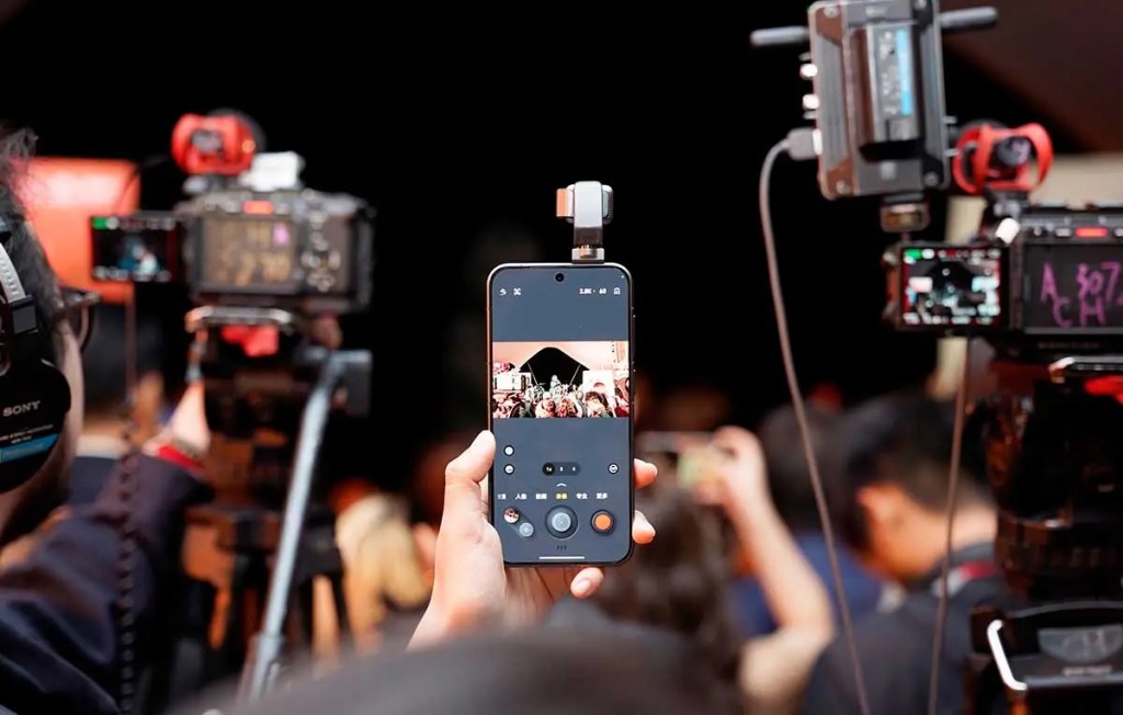 Una persona sosteniendo un teléfono móvil que muestra una imagen de un evento, rodeada de cámaras de grabación y un fondo de asistentes.