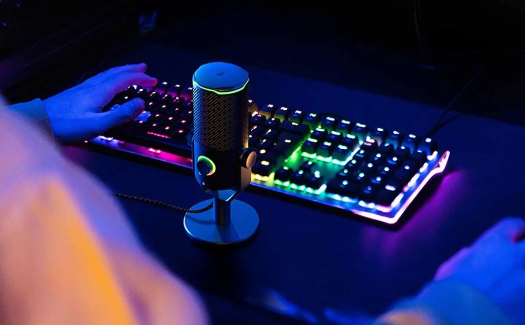 Un teclado retroiluminado con efectos de luz y un micrófono en un fondo oscuro, mientras una mano se apoya en el teclado.