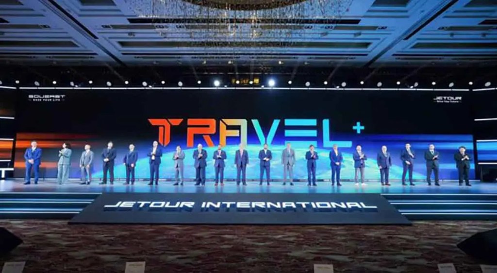 Grupo de personas en un escenario durante un evento de Jetour International, con un fondo iluminado que presenta las palabras 'TRAVEL+' y 'JETOUR INTERNATIONAL'.