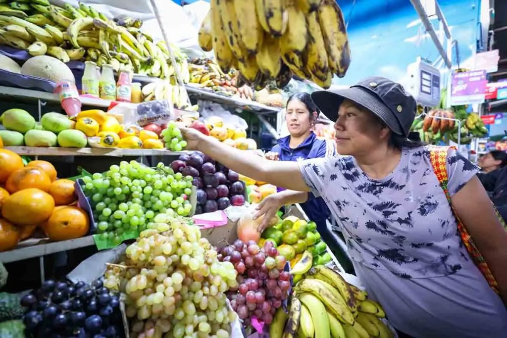 Mujer eligiendo frutas frescas en un mercado, rodeada de diversas opciones como uvas, plátanos y naranjas.