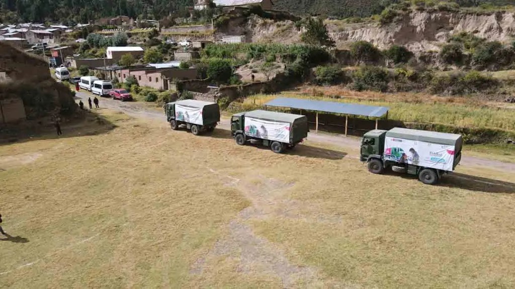 Tres camiones de carga en un campo, rodeados de un paisaje rural con casas pequeñas en el fondo.