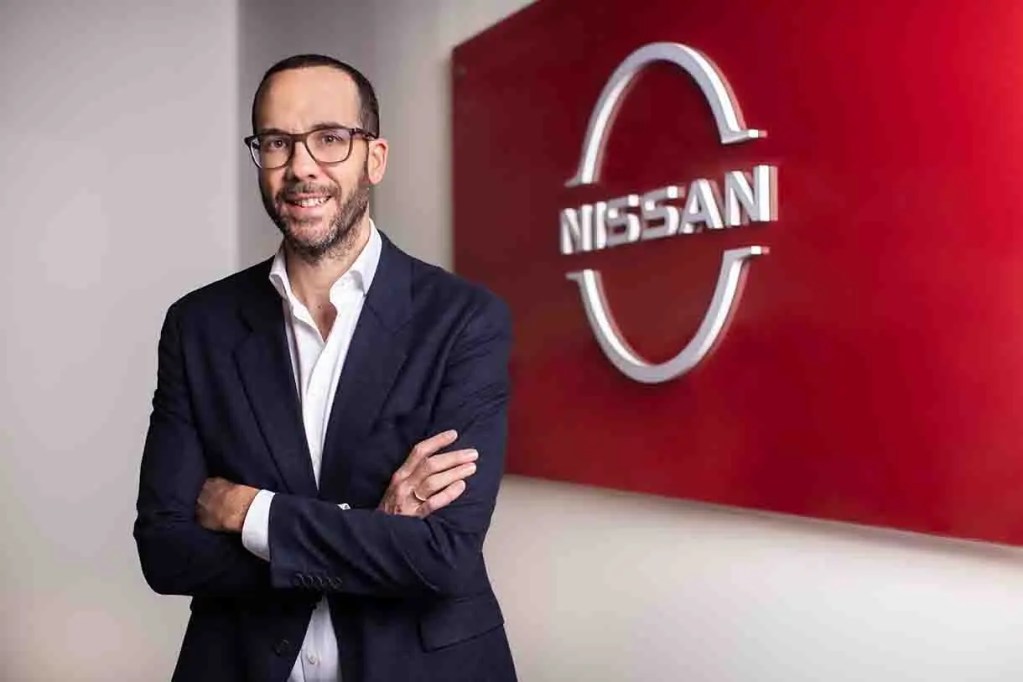 Hombre de pie sonriendo con brazos cruzados frente al logo de Nissan en una pared roja.