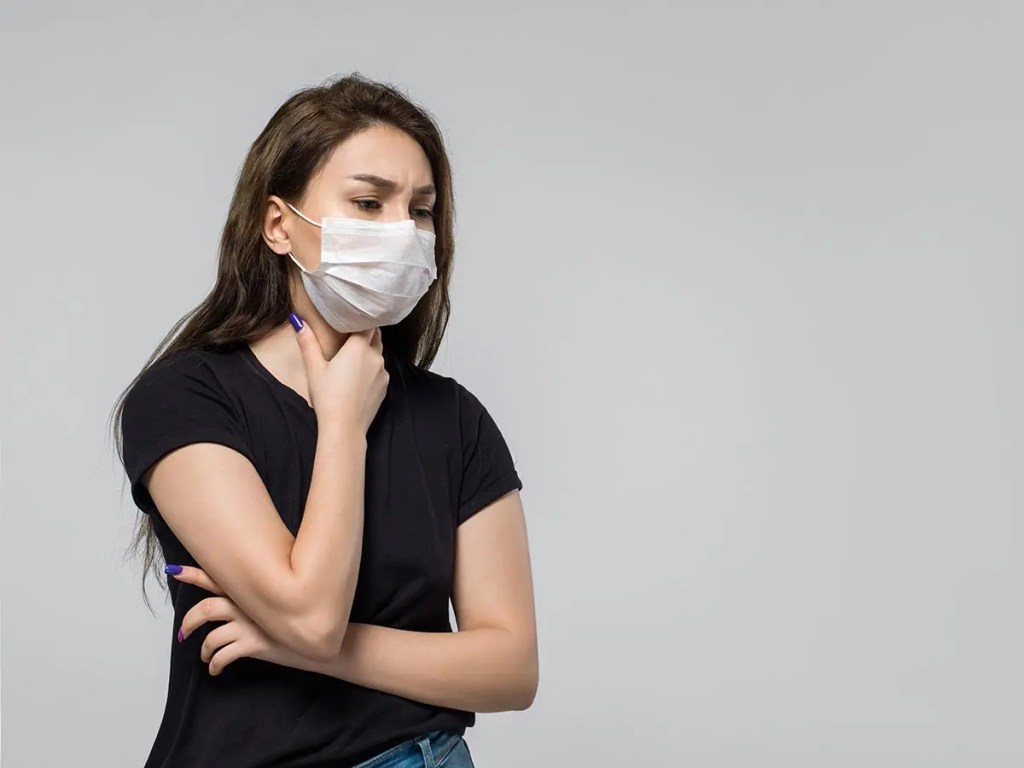 Mujer joven con mascarilla facial, mirando preocupada y tocándose la garganta, en un fondo gris.