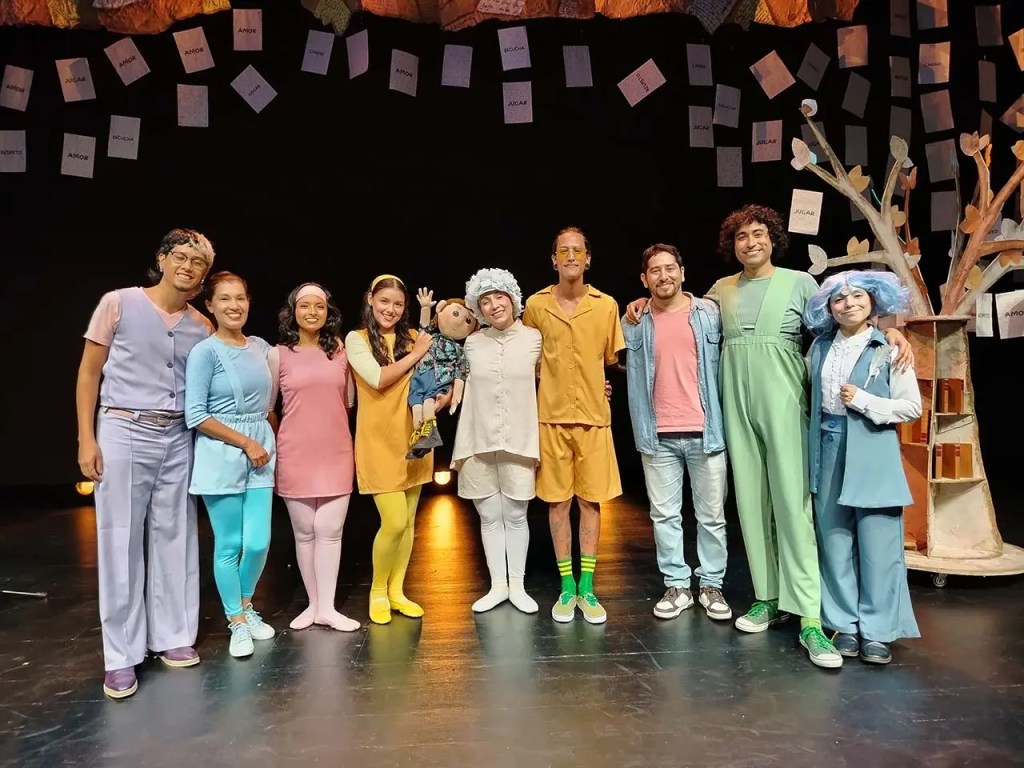Un grupo de ocho actores en un escenario, vestidos con disfraces coloridos, posando juntos después de una actuación teatral. En el fondo, se ven decoraciones y papeles colgantes.