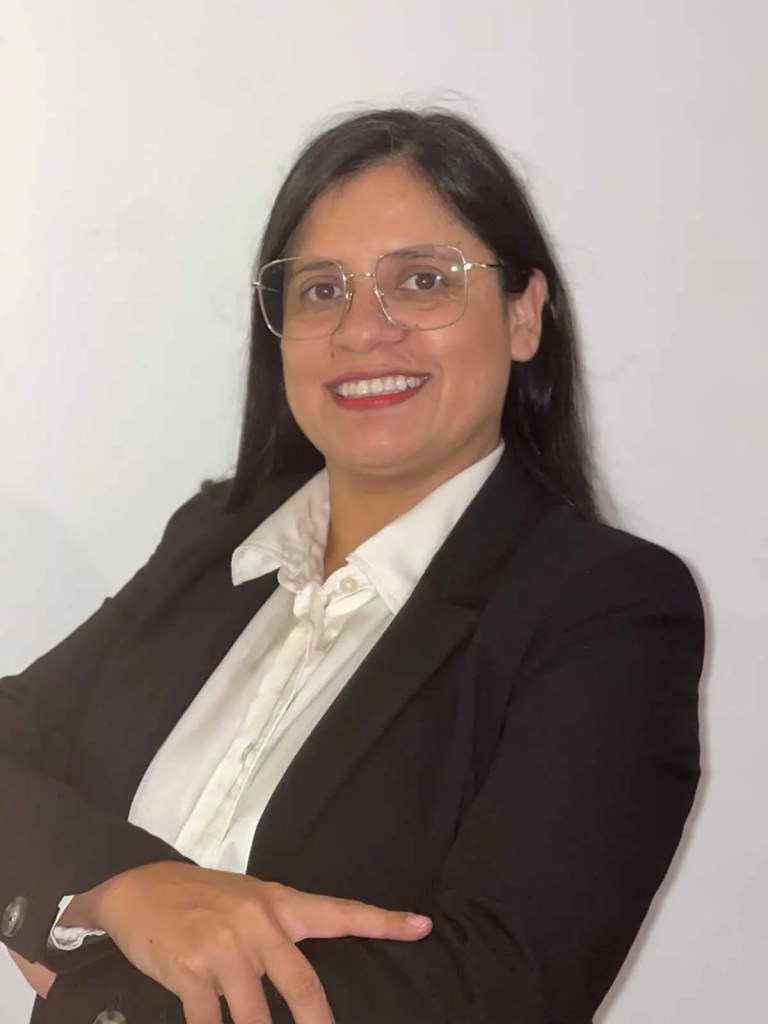 Mujer con cabello oscuro y gafas, vestida con chaqueta negra y camisa blanca, sonriendo y cruzando los brazos.