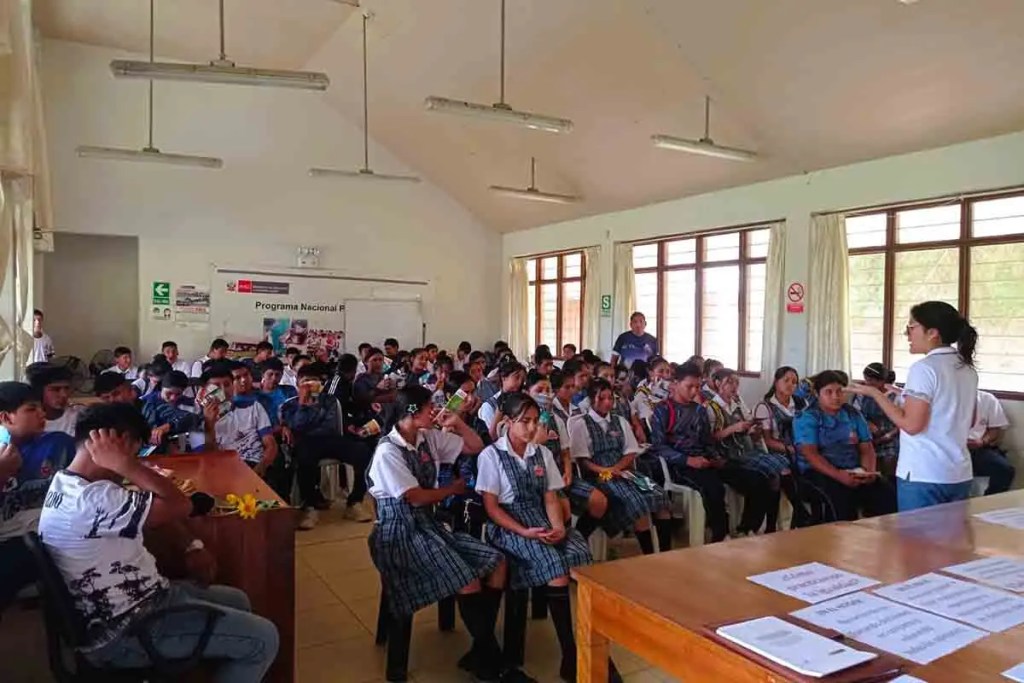 Un grupo de estudiantes sentados en un aula, escuchando a una presentadora que se encuentra de pie frente a ellos. El aula tiene ventanas grandes y está iluminada. Los estudiantes visten uniformes escolares.