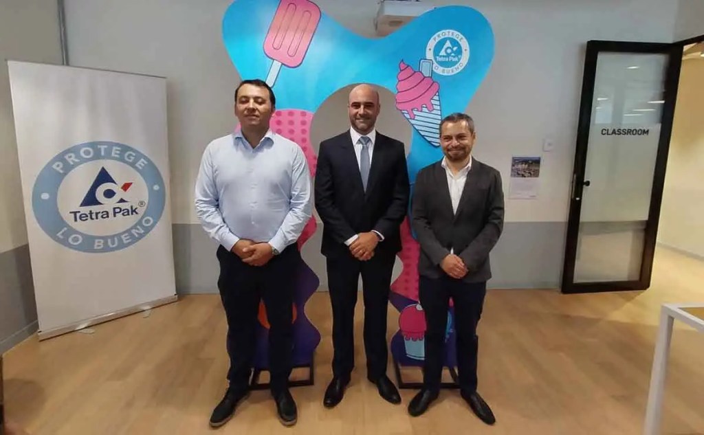 Tres hombres de pie frente a un fondo colorido con motivos de helados, junto a un cartel de Tetra Pak que dice 'Protege lo Bueno'.