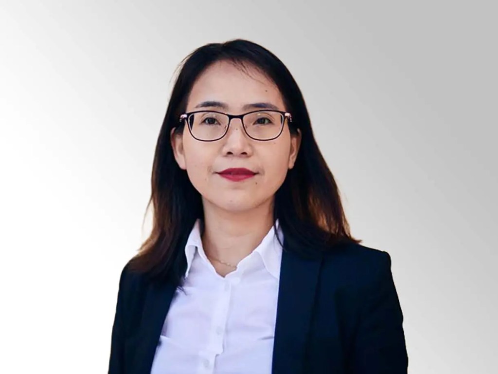 Mujer de pie con cabello largo y negro, usando gafas y un blazer negro sobre una camisa blanca, sonriendo frente a un fondo gris claro.