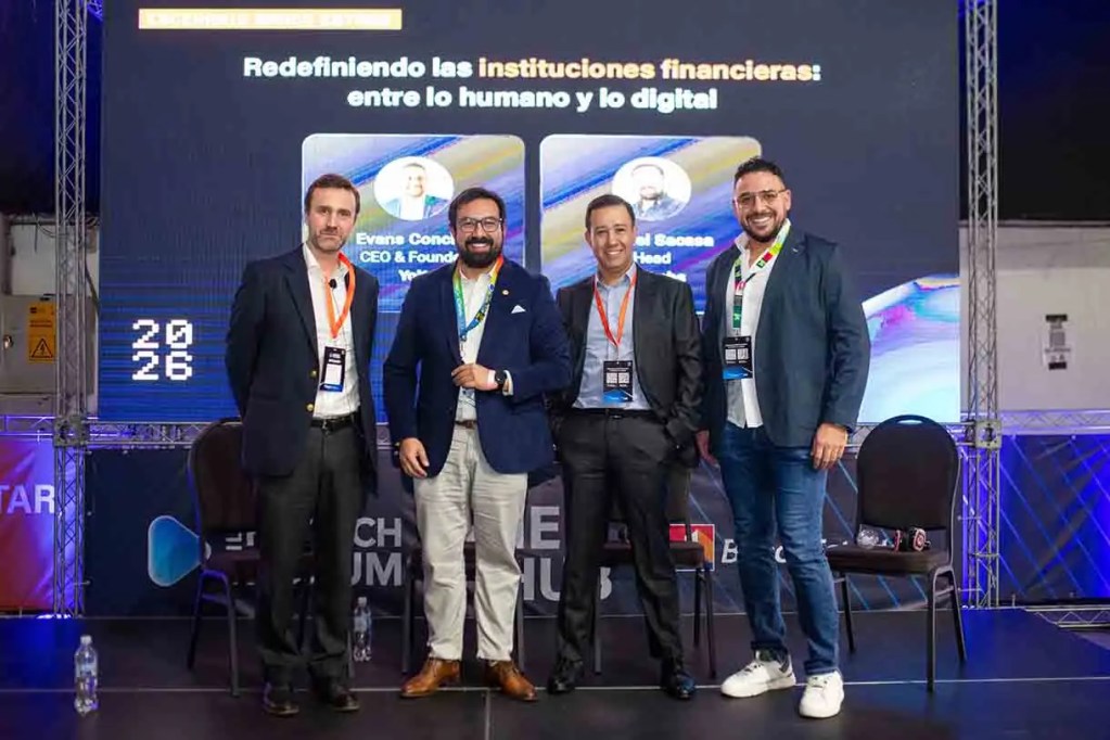 Cuatro ponentes en un evento sobre instituciones financieras, de pie y sonriendo frente a una pantalla con el título 'Redefiniendo las instituciones financieras: entre lo humano y lo digital'.