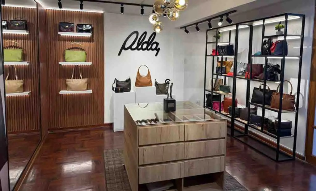 Tienda de accesorios con estantes que exhiben bolsos de varios colores y estilos, mostrador de madera en el centro, logo de la marca 'Aldo' en la pared.