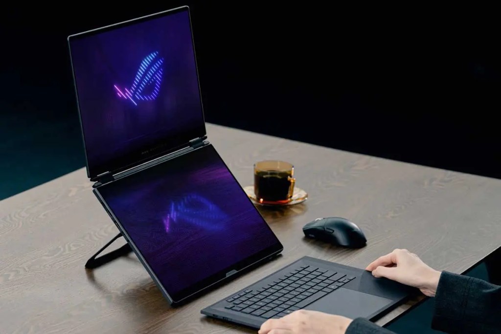 Una laptop con una pantalla doble, mostrando un diseño iluminado en colores oscuros, sobre una mesa de madera. Hay un mouse y una taza de café en la mesa, mientras una persona utiliza el teclado.