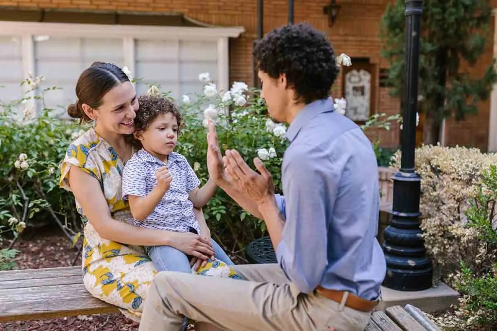 Una madre sonriente sentada en un banco con su hijo pequeño, mientras un hombre interactúa con ellos en un jardín con flores.