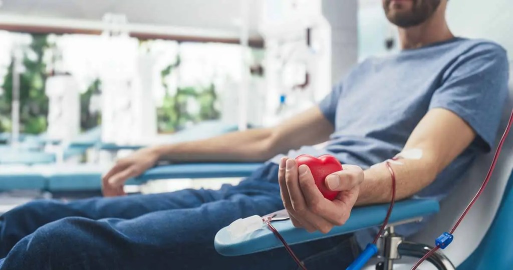 Un hombre sentado en una silla de donación de sangre, sosteniendo un corazón rojo de juguete con una aguja insertada en su brazo.