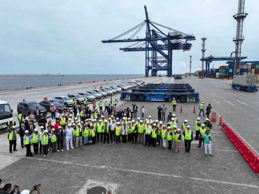 Grupo de personas en un puerto, con vehículos alineados y grúas en el fondo. Todos llevan chalecos reflectantes y algunos cascos de seguridad.