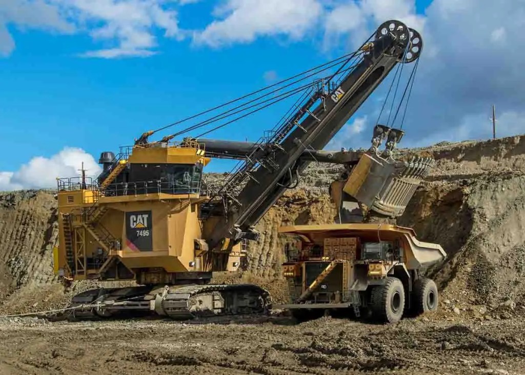 Excavadora de minería amarilla CAT 7495 trabajando en una cantera, levantando tierra con un camión volquete al lado.