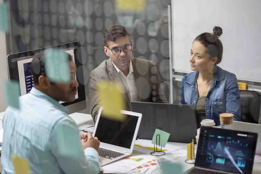 Reunión de trabajo con tres personas sentadas frente a computadoras, discutiendo gráficos y datos en un ambiente de oficina.