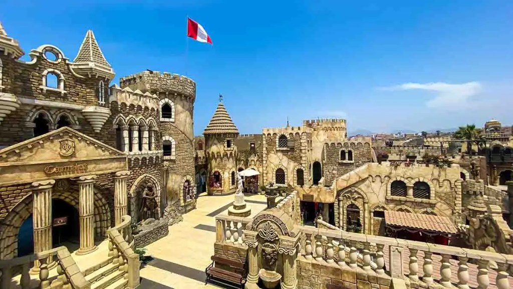 Vista panorámica de un complejo arquitectónico estilo medieval con torres y muros de piedra, adornado con una bandera peruana ondeando, bajo un cielo azul claro.